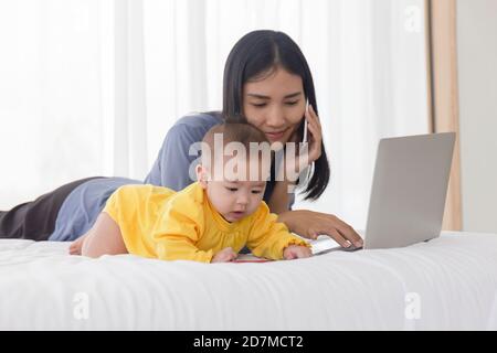 Ein asiatisches Baby spielt neben ihrer Mutter im Bett, während ihre Mutter am Handy spricht. Stockfoto