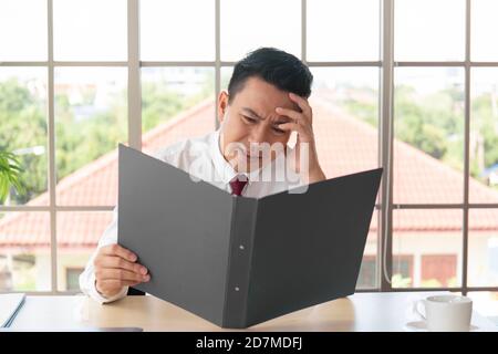 Der männliche Manager in Asien überprüfte das Firmenkonto und hatte ernsthafte Kopfschmerzen in seinem Büro. Stockfoto