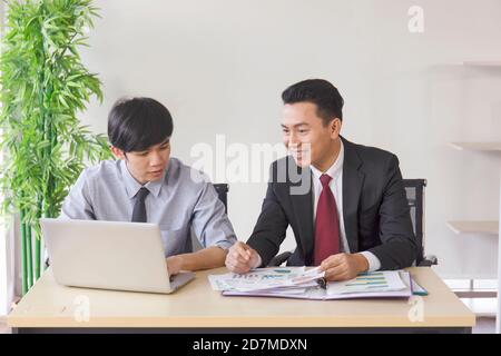 Ein asiatischer männlicher Supervisor bringt dem Neuling auf seinem Schreibtisch das Büro bei. Stockfoto