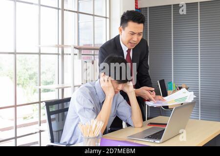 Ein wütender asiatischer männlicher Manager, der neben dem Schreibtisch eines Untergebenen steht und Dokumente hält. Stockfoto