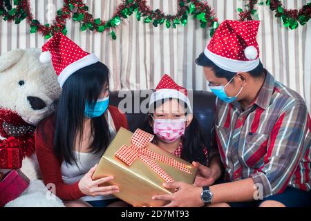 Glückliche Eltern geben Weihnachten Geschenk für Tochter mit tragen Gesicht Maske.Familie im weihnachtlichen Interieur Stockfoto
