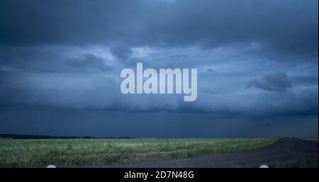 Dunkelblaue stürmische Wolken mit Blitzen schweben im Himmel über Gelbe Felder an brauner Bodenstraße gegen grüne Wald Zeitraffer Stockfoto