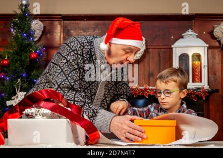 Glücklicher Großvater lehrt seinem Enkel, wie man Weihnachten einwickelt Geschenke auf dem Tisch Stockfoto