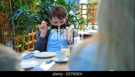 Schuljunge nimmt Gesichtsmaske aus und trinkt Saft aus Glas Sitzen im Restaurant mit langhaarigen Mutter Silhouette im Vordergrund Stockfoto