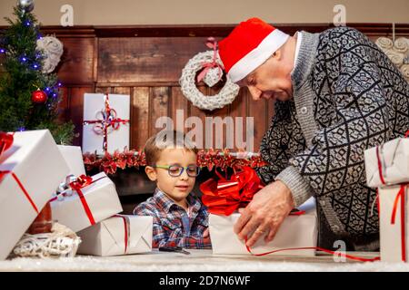 Glücklicher Großvater lehrt seinem Enkel, wie man Weihnachten einwickelt Geschenke auf dem Tisch Stockfoto