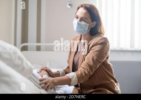 Junge Frau in Casualwear und Schutzmaske berühren Hände Kranker Freund Stockfoto