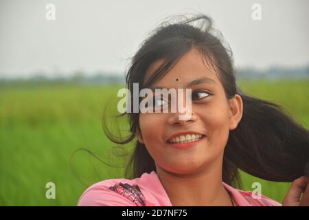 Nahaufnahme eines Teenagers in rosa Kleid, lange Haare, lächelnd und stehend auf einem Reisfeld mit grünen Pflanzen, selektive Fokussierung Stockfoto