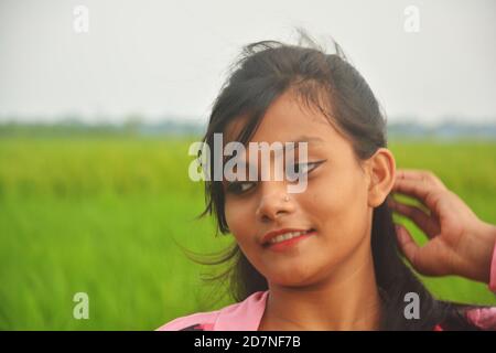 Nahaufnahme eines Teenagers mit rosa Kleid, langen Haaren, Händen auf dem Kopf, lächelnd und stehend auf einem Reisfeld mit grünen Pflanzen. Stockfoto