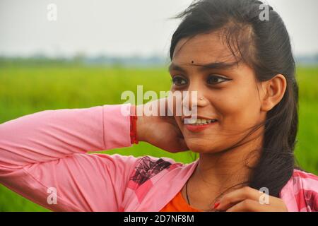 Nahaufnahme eines Teenagers mit rosa Kleid, langen Haaren, Händen auf dem Kopf, lächelnd und stehend auf einem Reisfeld mit grünen Pflanzen. Stockfoto