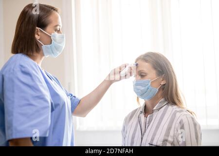 Junge Krankenschwester in blauer Uniform und Maske hält Thermometer durch Stirn des Patienten Stockfoto