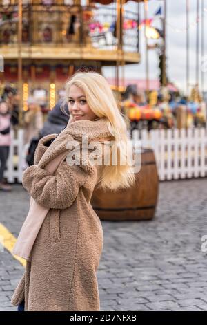01 14 2020 Moskau, Russische Föderation,schöne junge Frau in einem leichten Pelzmantel aus Kunstfell steht am Weihnachtskarussell und lächelt. Stockfoto