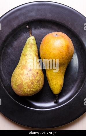 Zwei Birnenfrüchte, Birnen, Pyrus, flach auf dunkelschwarzem Rundteller auf Holztisch oder Schreibtisch, Food-Fotografie, Nahaufnahme, Kopierraum Stockfoto