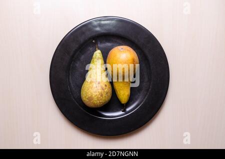 Zwei Birnenfrüchte, Birnen, Pyrus, flach auf dunkelschwarzem Rundteller auf Holztisch oder Schreibtisch, Food-Fotografie, Nahaufnahme, Kopierraum Stockfoto