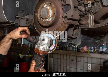Ein Mechaniker grinst, mit einem Winkelschleifer, die Kante einer alten hinteren Bremsscheibe in einem Auto, Regeneration des Bremssystems. Stockfoto