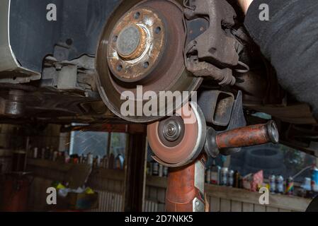 Ein Mechaniker grinst, mit einem Winkelschleifer, die Kante einer alten hinteren Bremsscheibe in einem Auto, Regeneration des Bremssystems. Stockfoto