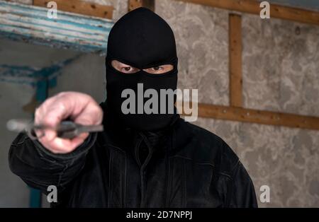 Ein Mann, der eine Maske/Kapuze in einem verlassenen Gebäude trägt. Er zeigt ein großes Messer auf den Betrachter. Nahaufnahme mit geringer Schärfentiefe. Stockfoto