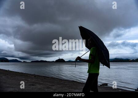 Ein Junge, der am 23. Oktober 2020 in Guwahati, Indien, einen Regenschirm am Ufer des Flusses Brahmaputra hält, während sich dunkle Wolken am Himmel sammeln. Stockfoto