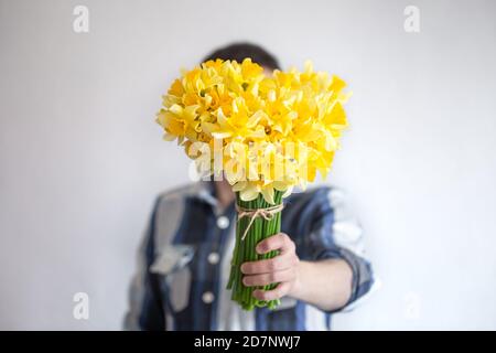 Ein Mann in einem Hemd bedeckt sein Gesicht mit einem Blumenstrauß. Auf hellem Hintergrund. Das Konzept der Grüße und Frauentag. Stockfoto