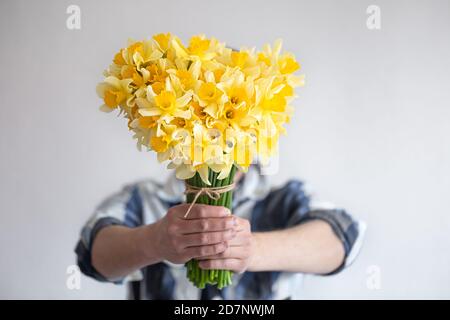 Ein Mann in einem Hemd bedeckt sein Gesicht mit einem Blumenstrauß. Auf hellem Hintergrund. Das Konzept der Grüße und Frauentag. Stockfoto