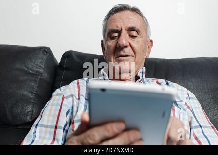 Ältere Männer in legerer Kleidung sitzen auf der Couch und browsen Tablet, während Freizeit zu Hause verbringen. Stockfoto