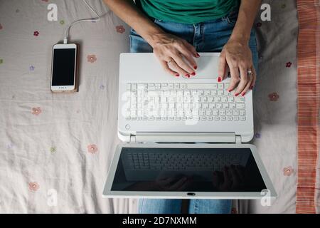 Von oben der Ernte anonyme Freiberuflerin auf dem Bett sitzend In der Nähe des Smartphones und beim Tippen von Informationen auf der Laptop-Tastatur während der Arbeit Entfernt von ho Stockfoto