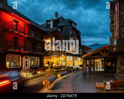 Abends Blick auf die Straße in einem kleinen Schweizer Dorf. Die hohen Alpen im Hintergrund scheinen gezeichnet zu sein. Warme Beleuchtung von gemütlichen Chalets. Stockfoto