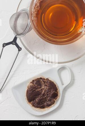 Gebrauchter Teebeutel auf Keramikplatte mit Glas Tee und Sieb Infuser auf weißem Hintergrund. Stockfoto