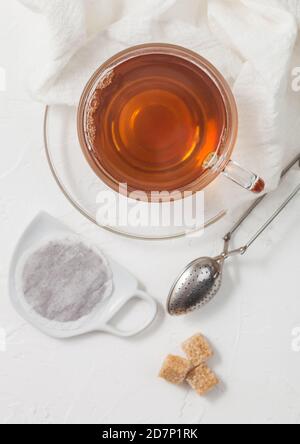 Schwarzer Tee-Teebeutel auf Keramikplatte mit Glas Tee und Sieb Infuser und Rohrzucker auf weißem Hintergrund. Stockfoto