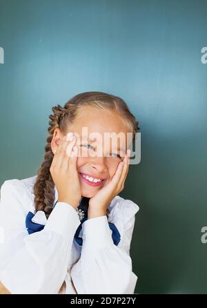 Ein Schulmädchen ein Mädchen mit schönen Zöpfen in einer Schule Uniform steht neben der Tafel und lächelt mit ihren Händen Auf ihrem Gesicht Stockfoto