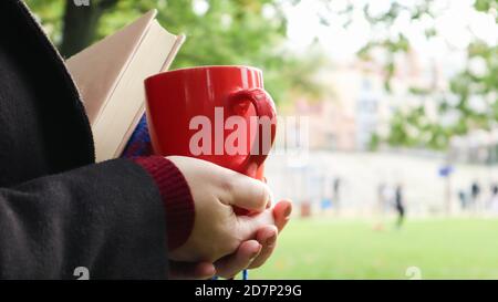 Eine rote Tasse Kaffee und ein Buch mit einer blau karierten Wolldecke oder einem karierten Tuch in den Händen einer Frau, die einen Pullover und einen schwarzen Mantel im Park trägt. W Stockfoto