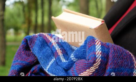 Ein Buch mit einer blau karierten Wolldecke oder einem Karomantel in den Händen einer Frau, die einen Pullover und schwarzen Mantel im Park trägt. Warmes und sonniges Wetter. Sof Stockfoto