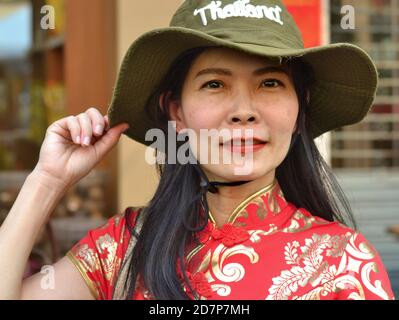 Die Thailändische Chinesin mittleren Alters trägt ein traditionelles chinesisches Kleid in Rot und Gold (Cheongsam) und einen grünen, breitkrempigen Thailand-Souvenir-Buschhut. Stockfoto