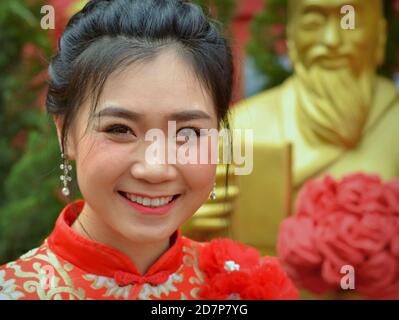 Schöne junge Thailänderin trägt ein traditionelles chinesisches Kleid in Rot und Gold (Cheongsam) und lächelt während des chinesischen Neujahrs für die Kamera. Stockfoto