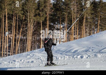 Glücklich lächelnder Mann in einem Skianzug klettert den Berg auf Skiern auf einer schneebedeckten Piste mit einem Schlepplift. Vor dem Hintergrund eines Nadelwaldes. Stockfoto