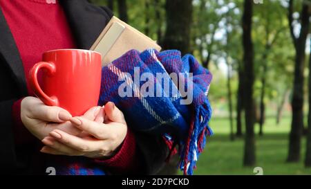 Eine rote Tasse Kaffee und ein Buch mit einer blau karierten Wolldecke oder einem karierten Tuch in den Händen einer Frau, die einen Pullover und einen schwarzen Mantel im Park trägt. W Stockfoto