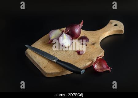 Das Messer und die Zwiebeln befinden sich auf einem Holzschneidebret. Rote Zwiebel, in Scheiben geschnitten und ganz. Schwarzer Hintergrund Stockfoto