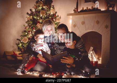 Familie mit zwei kleinen Jungen, die vor dem Weihnachtsbaum sitzen, liest der Vater nachts am Heiligabend eine Weihnachtsgeschichte Stockfoto