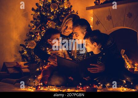 Familie mit zwei kleinen Jungen, die vor dem Weihnachtsbaum sitzen, liest der Vater nachts am Heiligabend eine Weihnachtsgeschichte Stockfoto