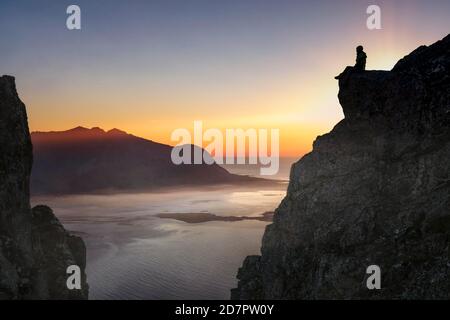 Silhouette einer Frau, die bei Sonnenuntergang auf einem Felsvorsprung sitzt, im Hintergrund Meer, Fjord und Berge mit Bodennebel, Gimsoy, Lofoten, Nordland, Norwegen Stockfoto