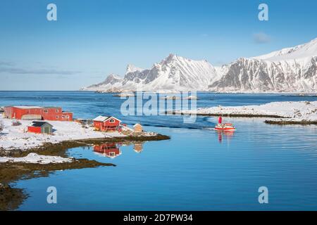 Rotes Bauernhaus und Fischerboot spiegeln sich im Fjord Rossoy Straumen, im Hintergrund das Meer, verschneite Berge Hustinden und Bjoerntinden Stockfoto