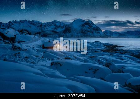 Tief verschneite Winterlandschaft am Meer, Holzhütte mit warmem Licht an der Küste, hinter Bergen unter Sternenhimmel, Kleppstad, Nordland, Lofoten Stockfoto