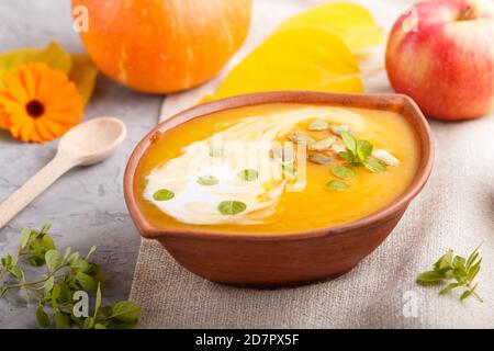 Traditionelle Kürbiscremesuppe mit Samen in Tonschüssel auf grauem Betongrund mit Leinentextilien. Seitenansicht, Nahaufnahme, selektiver Fokus. Stockfoto