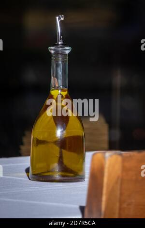 Eine große Glasflasche Olivenöl sitzt auf einem Restauranttisch mit einer weißen Baumwolltischdecke. Stockfoto