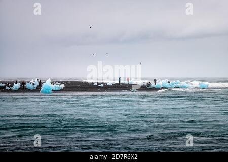 Wellen krachen auf blauen Gletschereisbergen Eis schwimmend und Menschen Touristen auf schwarzem Sand am Jokulsarlon Lagune See Diamant Strand In Island Stockfoto