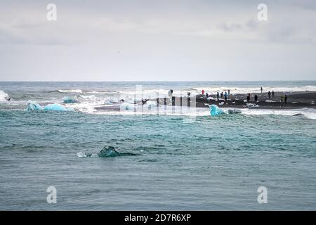 Wellen krachen auf blauen Gletschereisbergen schwimmend und Menschen Touristen Auf schwarzem Sand von Jokulsarlon Lagune See Diamant Strand in Island Stockfoto