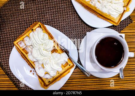 Closeup Flat Top Tisch Lay Blick auf geschmolzene heiße Schokolade Tasse und Löffel Untertasse, belgische Waffel auf weißem Teller mit Gabel mit Schlagsahne in mor gekrönt Stockfoto