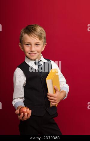 Portrait hübsch preteen Schuljunge in Uniform vor rotem Hintergrund. Netter Kerl halten gelbes Buch und Apfel. Tag des Wissens - 1. September Stockfoto