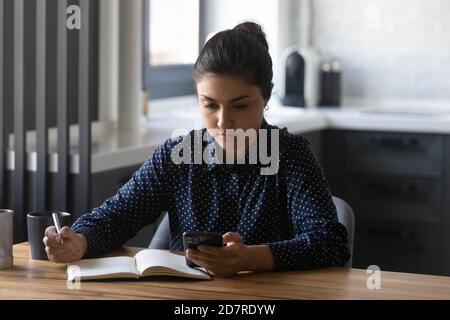 Fokussierte indische Frau schreiben in Notebook mit Smartphone Stockfoto