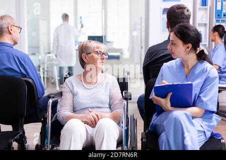 Krankenschwester, die Dokumente einreicht, während sie mit behinderten älteren Frauen im Wartebereich des Krankenhauses spricht. Patient fragt nach der Richtung an der Krankenhausaufnahme. Stockfoto