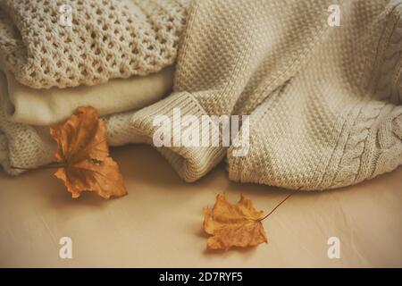 Auf einem beigen Hintergrund sind weiße Wollpullover warm gestrickt, und neben ihnen sind zwei trockene Ahornblätter. Warme Kleidung für kalte Herbsttage. Oktober. Stockfoto